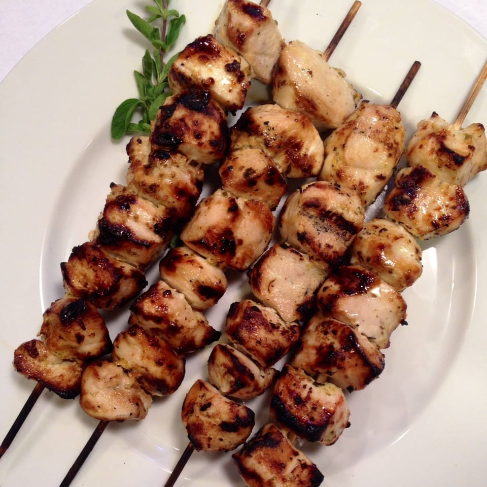 Three skewers of grilled chicken souvlaki with char marks and a sprig of oregano on a white plate.