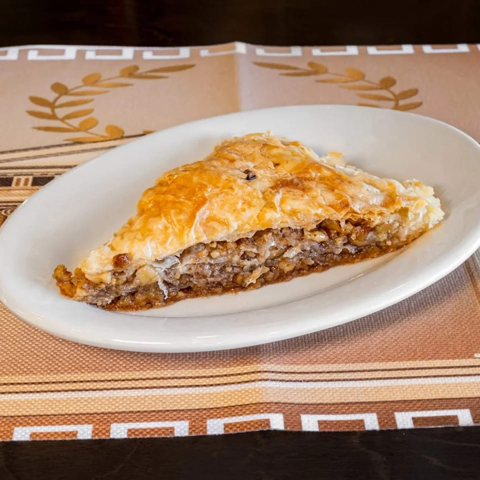 A triangular slice of golden-brown baklava with a rich nut filling on a white plate.