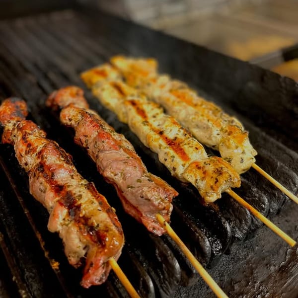 Four GRK Grill souvlaki skewers, two chicken and two pork, cooking on a hot grill.