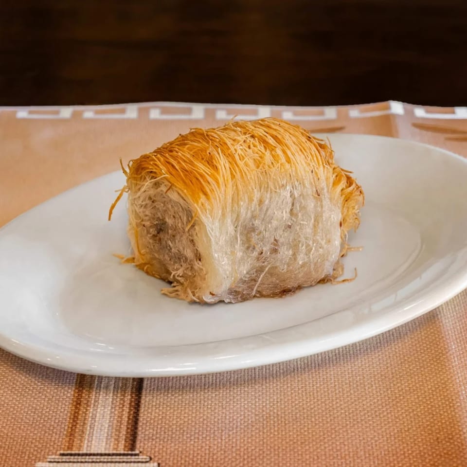 A single piece of golden-brown Greek kataifi pastry on a white plate, against a patterned tablecloth.